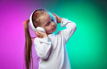 One beautiful little girl, kid listening music in headphones isolated on gradient purple blue color background in neon lightの写真素材