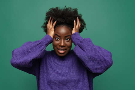 One beautiful girl holding her head with arms isolated on dark green studio background. Afro hairdo. Concept of emotions, facial expressionsの写真素材