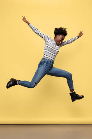 Full-length portrait of happy African girl in casual clothes jumping isolated on yellow studio...