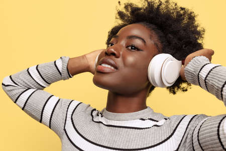 One attractive dark skinned girl in warm clothes in white headphones isolated on yellow background....