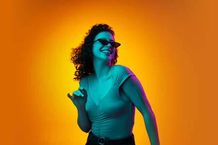 Portrait of young beautiful cheerful girl dancing isolated on yellow brown studio background in neon light.の写真素材