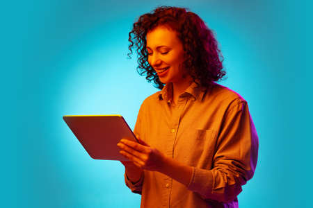 Studio shot of young pretty woman using tablet, gadget isolated on blue background in neon light.の写真素材