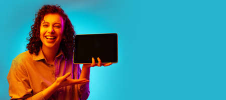 Studio shot of young pretty woman using tablet, gadget isolated on blue background in neon light.の写真素材