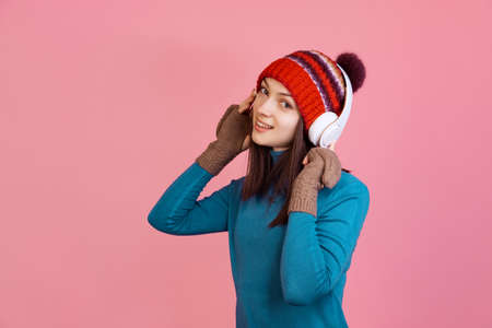 Young pretty girl in warm blue sweater, hat and winter mittens in headphones isolated on pink studio background.の写真素材