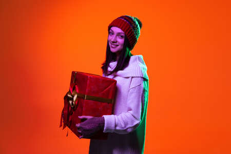 Studio shot of cute pretty young girl in warm clothes and knitted hat standing with gift box isolated on red background.の写真素材