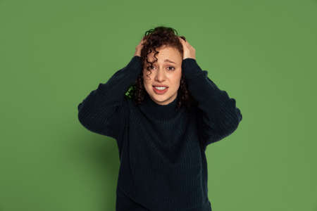One emotive young girl in warm winter sweater holding her head with arms isolated on green studio background. Concept of emotionsの写真素材