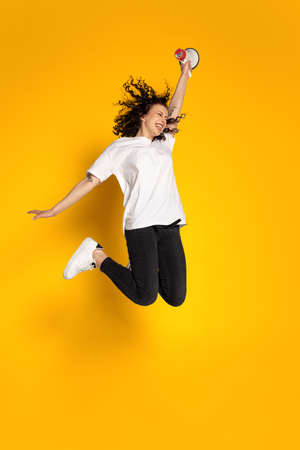 Full-length portrait of young cheerful girl, student in white t-shirt and jeans jumping isolated on yellow background.の写真素材