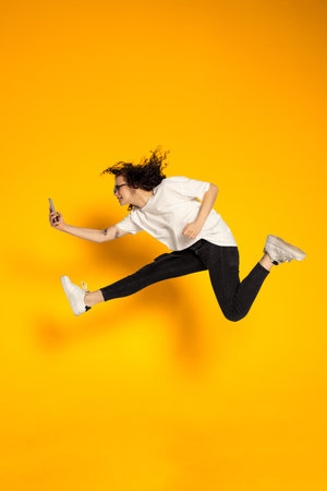 Full-length portrait of young cheerful girl, student in white t-shirt and jeans jumping isolated on yellow background.の写真素材