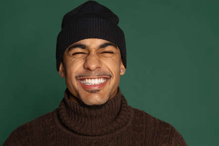 Close-up portrait of cheerful African young man in warm clothes and hat isolated on green color...