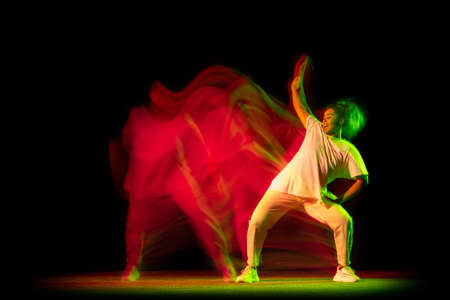 Studio shot of young active girl dancing hip-hop dance isolated on dark background in neon mixed yellow green light.の写真素材