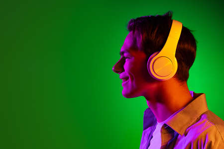 Portrait of young man, student listening to music in headphones isolated on green studio background...