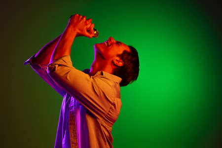 Portrait of young man, pop singer with microphone singing isolated on green studio background in...