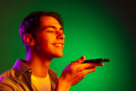 Smiling young man, student using gadget isolated on green studio background in neon light