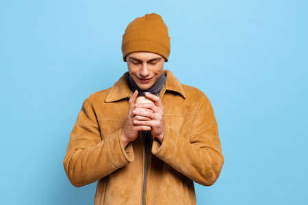 Young handsome happy man, student wearing warm winter clothes and hat drinking coffee on blue studio...