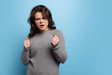 Studio shot of angry young beautiful girl in warm gray knitted sweater isolated on blue color backgroundの写真素材