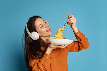 Cheerful young girl tasting delicious Italian pasta isolated on blue studio background. World pasta...
