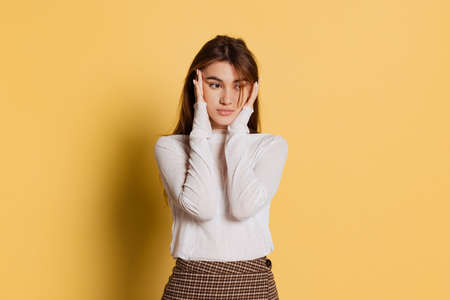 Sad, serious young beautiful girl in warm white neck-polo posing isolated on yellow color...