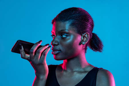 Portrait of young beautiful dark skinned woman using phone isolated on blue studio background in neon light.の写真素材