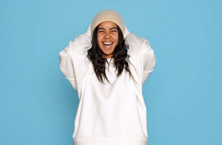 Happy, excited young beautiful dark skinned girl in warm white hoodie and hat isolated on blue...