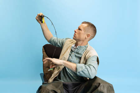 Serious young man, fisherman with fishing accessories sitting on chair isolated over blue studio...