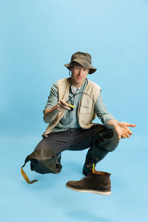 Funny young man, fisherman with fishing accessories wearing sport style clothes isolated over blue...