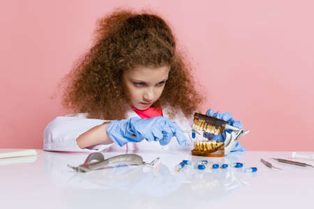 Comic portrait of little girl, child in image of dentist doctor wearing white lab coat and gloves isolated on pink studio backgroundの写真素材