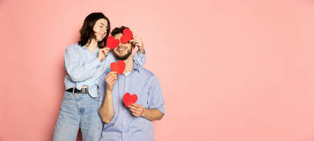 Flyer. Young and happy man and woman holding greeting cards shaped hearts isolated on pink trendy color background. Emotions, youth, love and lifestyle conceptの写真素材