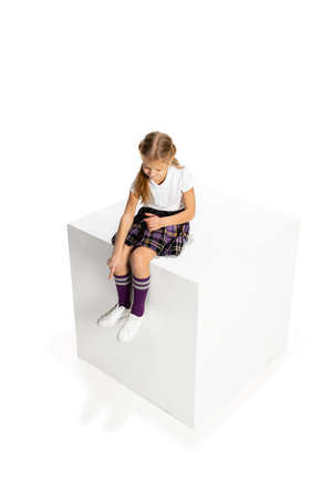 Studio shot of beautiful little girl, pupil in school uniform sitting on big box isolated on white background. Concept of childhood, emotions, studyの写真素材