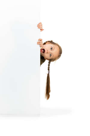 Funny child, little girl, pupil in casual style clothes peeking out from behind wall isolated on white background. Concept of childhood, emotions, studyの写真素材