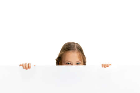 Funny child, little girl, pupil in casual style clothes peeking out from behind wall isolated on white background. Concept of childhood, emotions, studyの写真素材