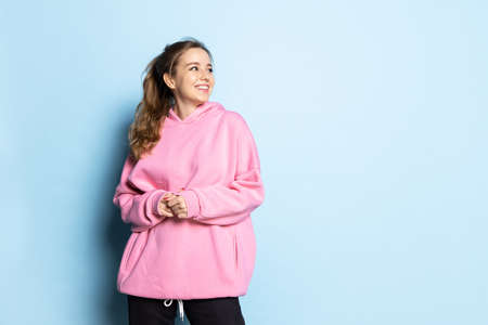 Half-length portrait of young beautiful girl wearing pink hoodie isolated on blue background. Concept of emotions, facial expression, youth, aspiration, salesの写真素材