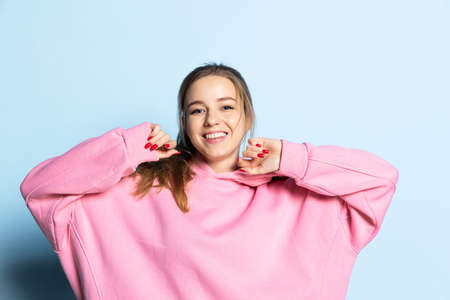 Half-length portrait of young beautiful girl wearing pink hoodie isolated on blue background. Concept of emotions, facial expression, youth, aspiration, salesの写真素材