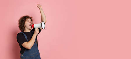 Flyer with portrait of young hairy man, student in black t-shirt shouting at megaphone isolated on pink background.の写真素材