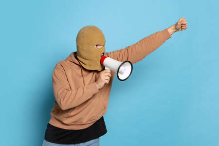 Studio shot of young anonymous man, student wearing balaclava shouting at megaphone isolated on blue...