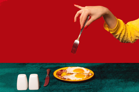 Human hand tasting bacon and eggs isolated on green and red background. Vintage, retro style interior. Food pop art photography.の写真素材