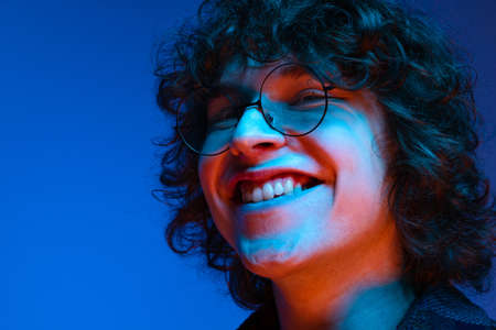 Portrait of young smiling man, student in black hoodie isolated on dark blue background in neon...