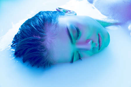 Close-up female face, young beautiful girl in milk bath with soft glowing in blue-green neon light....