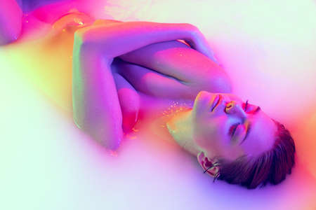 Portrait of attractive young girl in milk bath with soft glowing in white-pink neon light. Copyspace...