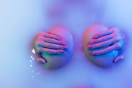 Close-up womans breasts closed by female hands isolated in milk liquid with soft glow in blue-pink...