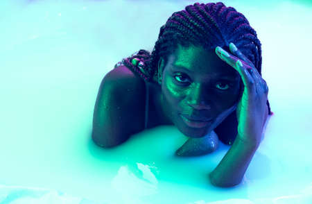 Portrait of beautiful young dark skinned girl lying in milk bath with soft glow in blue-pink neon light. Natural beauty, fashion, style, skincare conceptの写真素材