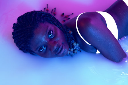 Portrait of beautiful young dark skinned girl lying in milk bath with soft glow in blue-pink neon...