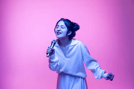 Portrait of young beautiful smiling girl in white blouse singing at microphone isolated on pink background in neon. Concept of emotionsの写真素材