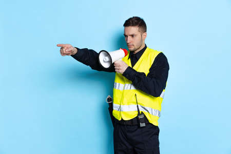 Portrait of young policeman officer wearing black uniform and warnvest shouting at megaphone isolated on blue background.の写真素材