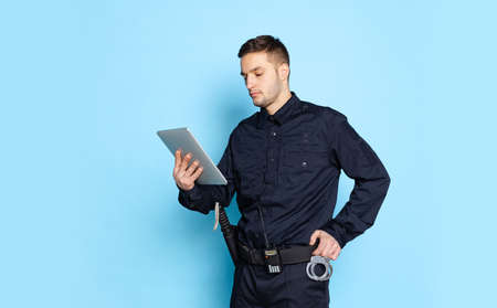 Portrait of young man, policeman officer wearing black uniform using tablet isolated on blue background.の写真素材