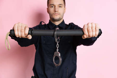One handsome policeman officer wearing black uniform with club and handcuffs isolated on pink background.の写真素材
