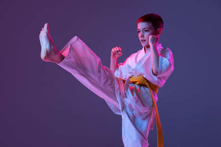 Portrait of sportive kid, male taekwondo, karate athletes in doboks doing basic movements isolated on purple background in neon. Concept of sport, martial artsの写真素材
