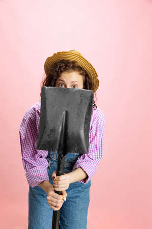 Comic portrait of young scared girl, gardener in work uniform and straw hat hiding behind shovel isolated on pink background. Concept of job, emotions, agronomy, funの写真素材