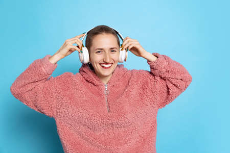 Happy pretty girl, student with short hair wearing pink hoodie in headphones isolated on blue...