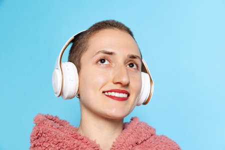 Happy pretty girl, student with short hair wearing pink hoodie in headphones isolated on blue...