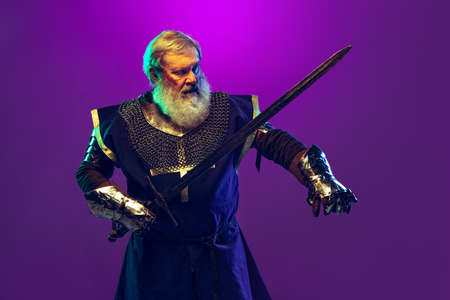 Portrait of senior greybearded man, brave and brutal medieval warrior or knight in armor with sword isolated over purple background in neon. Comparison of erasの写真素材